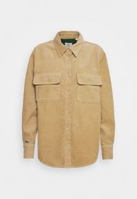 Camisa de corduroy en color beige claro, con dos bolsillos en el pecho, cierre de botones y cuello. Tejido texturizado con un patrón acanalado.