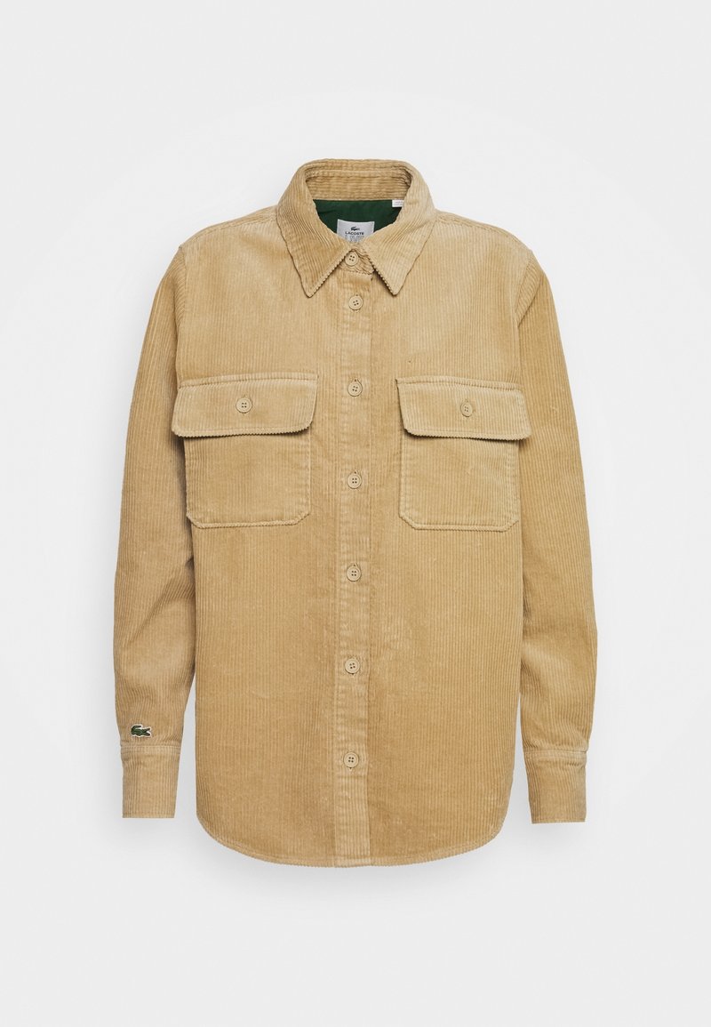 Camisa de corduroy en color beige claro, con dos bolsillos en el pecho, cierre de botones y cuello. Tejido texturizado con un patrón acanalado.