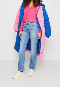 Abrigo acolchado azul y rosa con capucha, combinado con una camiseta rosa y jeans rectos de un azul claro. Zapatillas blancas completan el atuendo.