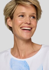 Weißes T-Shirt mit kurzen Ärmeln und blauem abstraktem Design auf der Vorderseite, runder Halsausschnitt und weicher Baumwollqualität. Das Model lächelt.