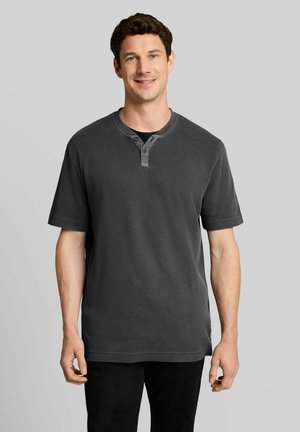 Uomo sorridente che indossa una maglietta Henley a maniche corte grigio scuro, in piedi davanti a uno sfondo grigio chiaro uniforme.