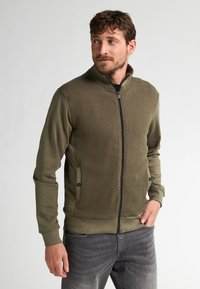 Olivengroen zip-up jack met een textuur aan de voorzijde, gladde mouwen, ritszakken en geribbelde manchetten. Het ontwerp heeft een hoge kraag en een getailleerde vorm.