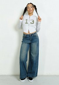 Szara, krótka bluza z kapturem i zamkiem, z zielonym graficznym napisem "13", zestawiona z szerokimi, niebieskimi dżinsami i czarnymi sneakersami. Gładkie faktury, luźny krój.