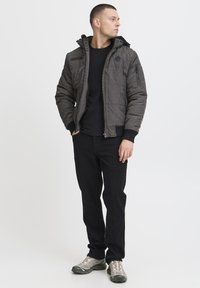 Gray quilted jacket with a hood, featuring a zip front, multiple pockets, and ribbed cuffs, paired with a black shirt and black pants.
