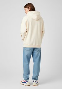 Sudadera crema con capucha, que cuenta con un detalle gráfico en la manga. Combinada con jeans azul claro y zapatillas blancas con acentos rojos y azules.