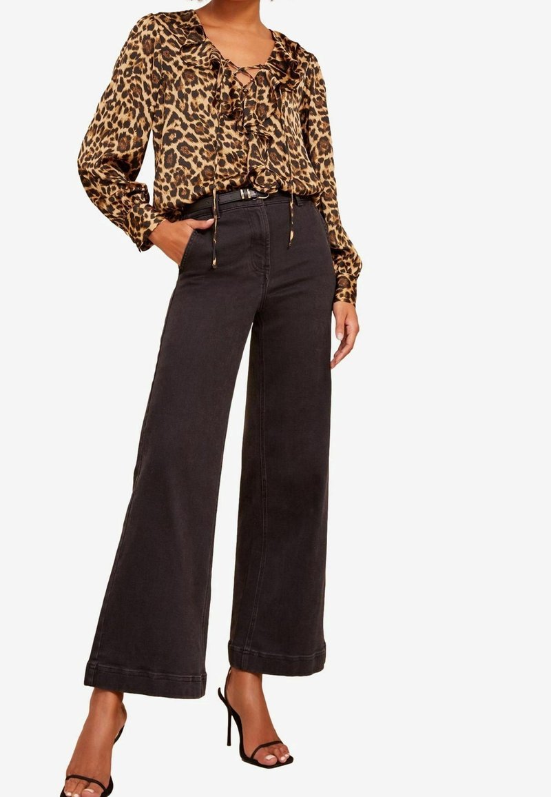 Femme portant un chemisier à manches longues imprimé léopard avec des volants et un pantalon noir taille haute à jambes larges, accompagné de talons noirs à brides.