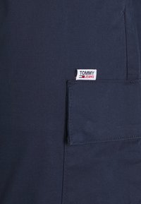 Námořnicky modrá tkanina s detailem kapsy; obsahuje malou bílou štítek s červenými a modrými akcenty s nápisem "TOMMY JEANS." Hladká textura.