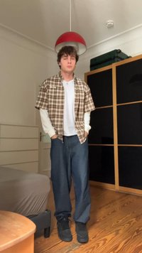 Karierte Kurzarmbluse über einem weißen Langarmshirt, kombiniert mit lockeren blauen Jeans und schwarzen Turnschuhen, steht in einem Raum mit Holzboden.