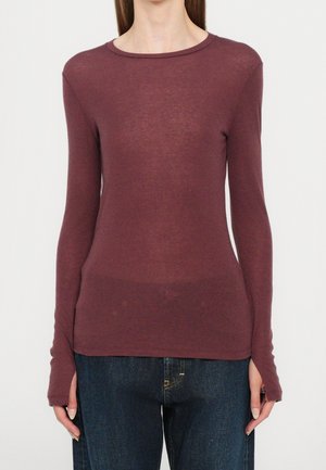 Femme portant un haut ajusté à manches longues couleur prune, associé à un jean bleu foncé, debout devant un fond uni.