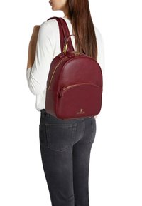 Zaino in pelle bordeaux con parte superiore arrotondata, dettagli in oro e tasca frontale con cerniera. Particolari includono il logo sulla parte bassa frontale.