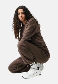 Bruine sweatshirt en joggingsbroek van een zachte stof, met een relaxed fit. Witte sportschoenen met zwarte en zilveren accenten. Krullend haar zichtbaar.