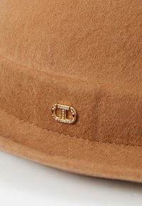 Brūns filca cepure ar plašu malām un zelta krāsas logotipa akcentu ar rhinestones. Gluda tekstūra un vienmērīga krāsa visā virsmā.