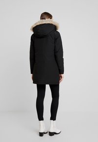Černá parka s kapucí lemovanou kožešinou, která má elegantní design a střední délku. Modelka má na sobě černé legíny a bílé kotníkové boty.