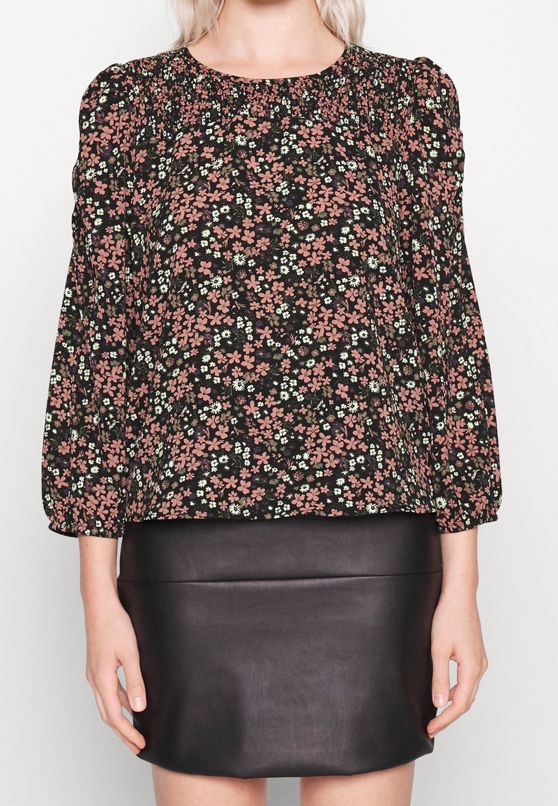 Blouse noire à motifs floraux avec des fleurs roses et vertes, présentant un détail froncé autour de l'encolure et des manches longues bouffantes.