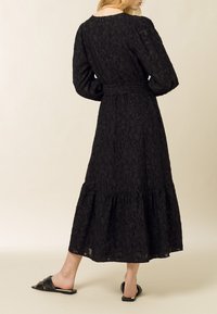 Robe noire à manches longues avec un motif floral texturé, présentant une taille cintrée, une jupe à volants et des détails plissés ; associée à des sandales noires.