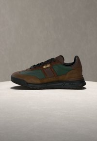 Hnědé a zelené tenisky z semišového a síťového materiálu, s černou gumovou podrážkou, zlatým logem a kontrastními pruhy na boku.