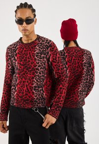 Suéter de leopardo en rojo y negro, con textura de punto, cuello redondo, combinado con jeans negros desgastados y un gorro de punto rojo.