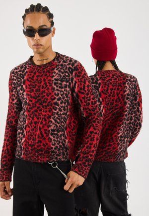 Maglione con stampa leopardata rossa e nera, texture a maglia, scollatura a girocollo, abbinato a jeans neri strappati e un berretto a maglia rosso.