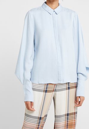 Person iført lyseblå bluse med lange ærmer og strukturerede manchetter samt beige, sorte og blå ternede bukser, stående mod en ensfarvet baggrund.