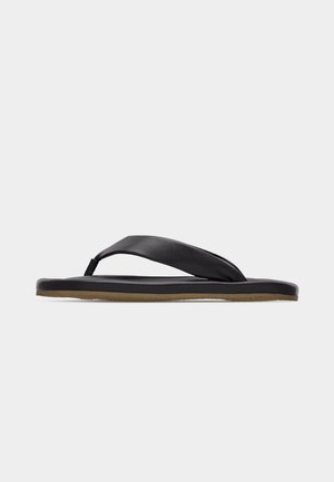 Melna pludmales čība ar mīkstinātu Y formas siksnu un plakanu zolīti uz vienkārša gaiša fona, sānskats.