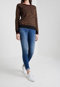 Bruin leopardenprint trui met zwarte randen, gecombineerd met slanke blauwe jeans en witte sneakers, met omgeslagen mouwen en een casual pasvorm.