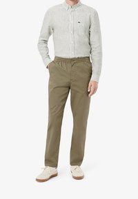 Osoba stojąca ubrana w zieloną i białą koszulę na guziki w paski, spodnie khaki z elastycznym pasem oraz kremowe trampki z brązowymi podeszwami.