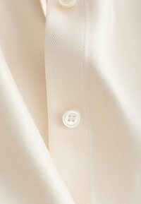 Gros plan sur une chemise couleur crème avec une texture sergée en diagonale, des boutons blancs et des coutures visibles sur le tissu.