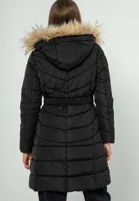 Abrigo inflable negro con capucha de pelo sintético, cintura ceñida con cinturón y detalle de costura en chevron. Textura suave, diseño de longitud media y mangas largas.