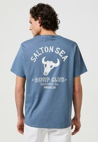 Camiseta de algodón azul con un gráfico blanco de un cráneo de vaca, texto "SALTON SEA SURF CLUB" y logo "WRANGLER" en la parte trasera. Mangas cortas.