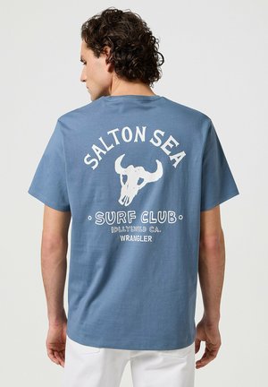 Blå bomullströja med ett vitt motiv av ett nötkreatursskelet, texten "SALTON SEA SURF CLUB" och "WRANGLER"-logotypen på baksidan. Kort ärm.