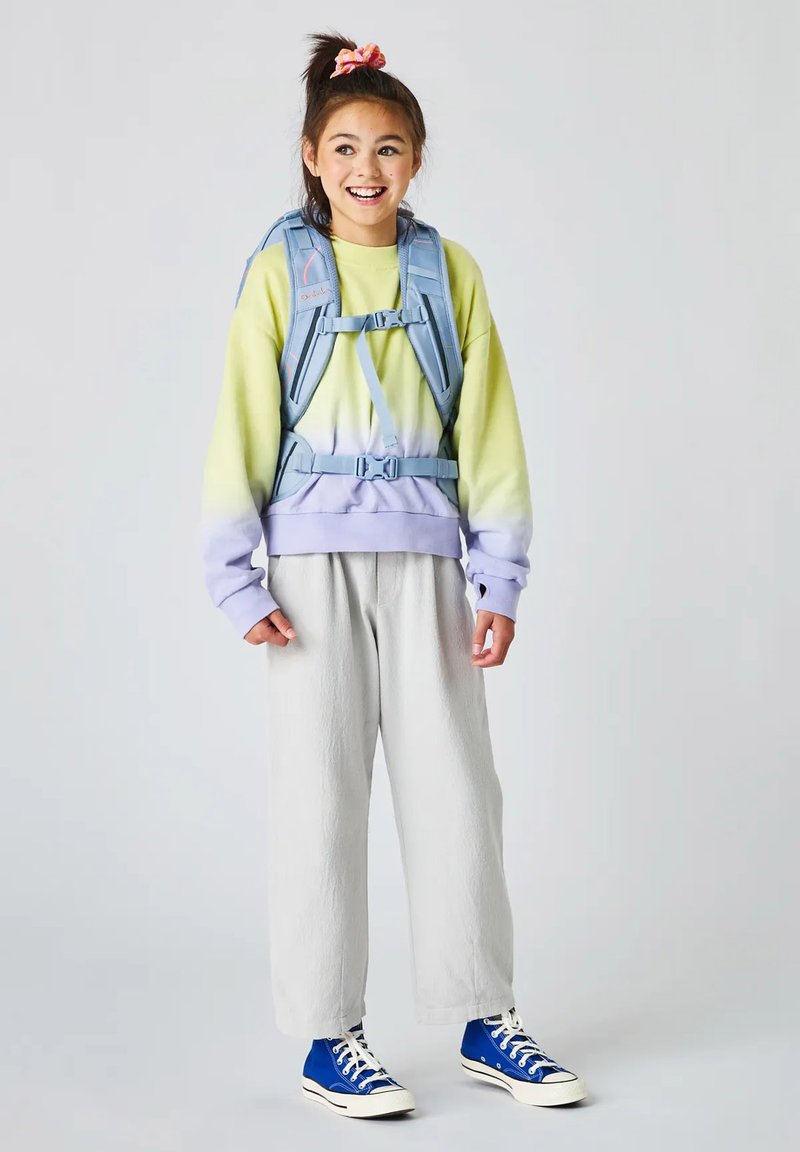 Lächelndes Mädchen, das einen pastellfarbenen Pullover, helle Hosen, blaue High-Top-Sneakers und einen hellblauen Rucksack mit Brust- und Taillengurten trägt.