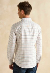 Biela košeľa s dlhými rukávmi a farebnou mriežkovou potlačou v červenej a modrej farbe. Má golier s gombíkmi a jemné logo na chrbte.