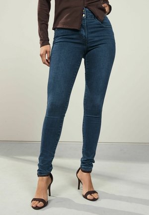 Schmal geschnittene dunkelblaue Jeans, kombiniert mit braunen offenen High Heels und einem braunen Langarmshirt, von der Taille bis zu den Füßen vor einfarbigem Hintergrund gezeigt.