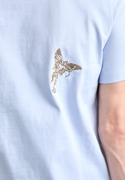 Ljusblå bomulls t-shirt med guldbroderad fjärilsmotiv på ryggen, med korta ärmar och en mjuk textur.