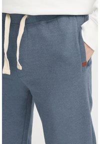 Pantalones de chándal azules hechos de una tela suave con cintura ajustable y bolsillos laterales. Presenta un pequeño parche de logo marrón en el lado izquierdo.