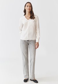 Blusa a maniche lunghe crema chiaro con scollatura a V e dettagli in pizzo sul collo, abbinata a jeans grigi chiari a gamba dritta e ballerine nere.