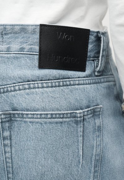 Ανοιχτό μπλε τζιν με μαύρο δερμάτινο ταμπελάκι με την επιγραφή "Won Hundred" στη μέση, φορεμένο με λευκό τοπ.