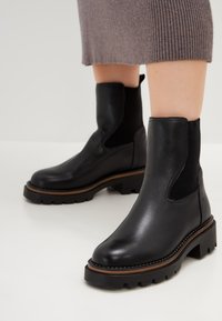 Botas de cuero negro hasta el tobillo con paneles elásticos laterales, suela gruesa y ribete marrón en contraste; textura suave con un diseño elegante.