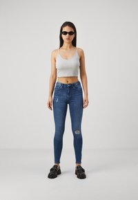 Top corto gris, jeans ajustados azules con una rodillera rasgada y zapatos de plataforma negros. Modelo usando gafas de sol, de pie contra un fondo neutral.
