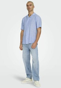 Korte mouwen, lichtblauw overhemd met knopen in een zachte, gestructureerde stof, gecombineerd met loszittende lichtblauwe jeans en witte sneakers.