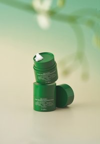 Zielone cylindryczne słoiki z balsamem oczyszczającym ułożone jeden na drugim, z jednym zdjętym wieczkiem, które odsłania białą substancję. Na boku znajdują się fakturowane etykiety z informacjami o produkcie.