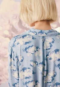 Ljusblå blus med ett molnmönster i vitt och marinblått, med mjuk textur, en krage och knappdetalj längs ryggen.