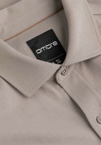 Chemise beige à col plié avec une étiquette noire visible inscrite "ombre" en blanc, taille EUR 48, avec deux boutons près du col.