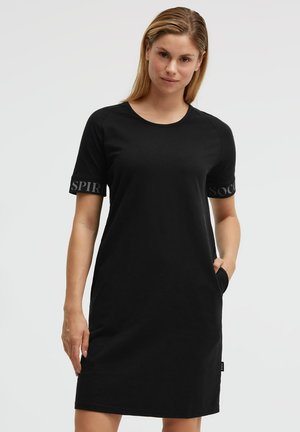 Frau trägt ein schwarzes, knielanges Kleid mit kurzen Ärmeln und Text auf den Ärmeln, eine Hand in der Tasche, steht vor einem schlichten hellen Hintergrund.