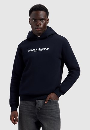 Jonge man met een donkere huid die een marineblauwe hoodie draagt met de tekst "BALLIN'", staand met één hand in de zak, neutrale grijze achtergrond.