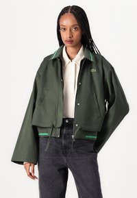 Groene cropped jas met een ritsluiting, gelaagde kraag met groene accenten en zijzakken, gedragen over een lichtgekleurde polo.