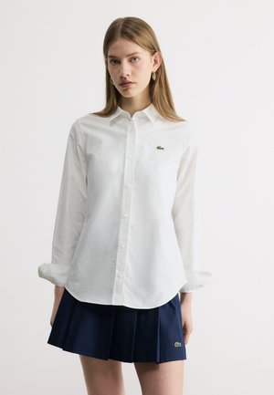 Sirge juuksega naine kannab valget nööpidega särki ja tumesinist voltidega seelikut, mõlemal vasakul küljel väike roheline krokodillilogoga.