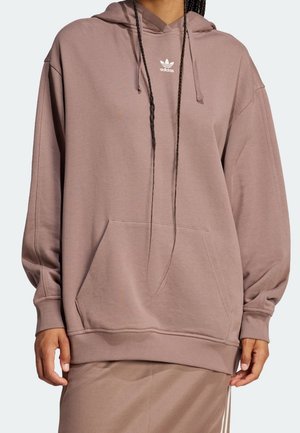 Personne portant un sweat à capuche Adidas mauve surdimensionné avec poche kangourou à l'avant et logo sur la poitrine, assorti à un bas assorti.