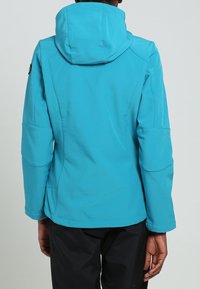 Personne portant une veste à capuche bleu vif et un pantalon noir, vue de dos sur un fond gris clair uni.