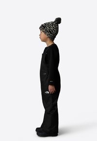 Black waterproof overalls with shoulder straps, paired with a knit hat featuring a zebra pattern and a pom-pom. Black footwear visible.